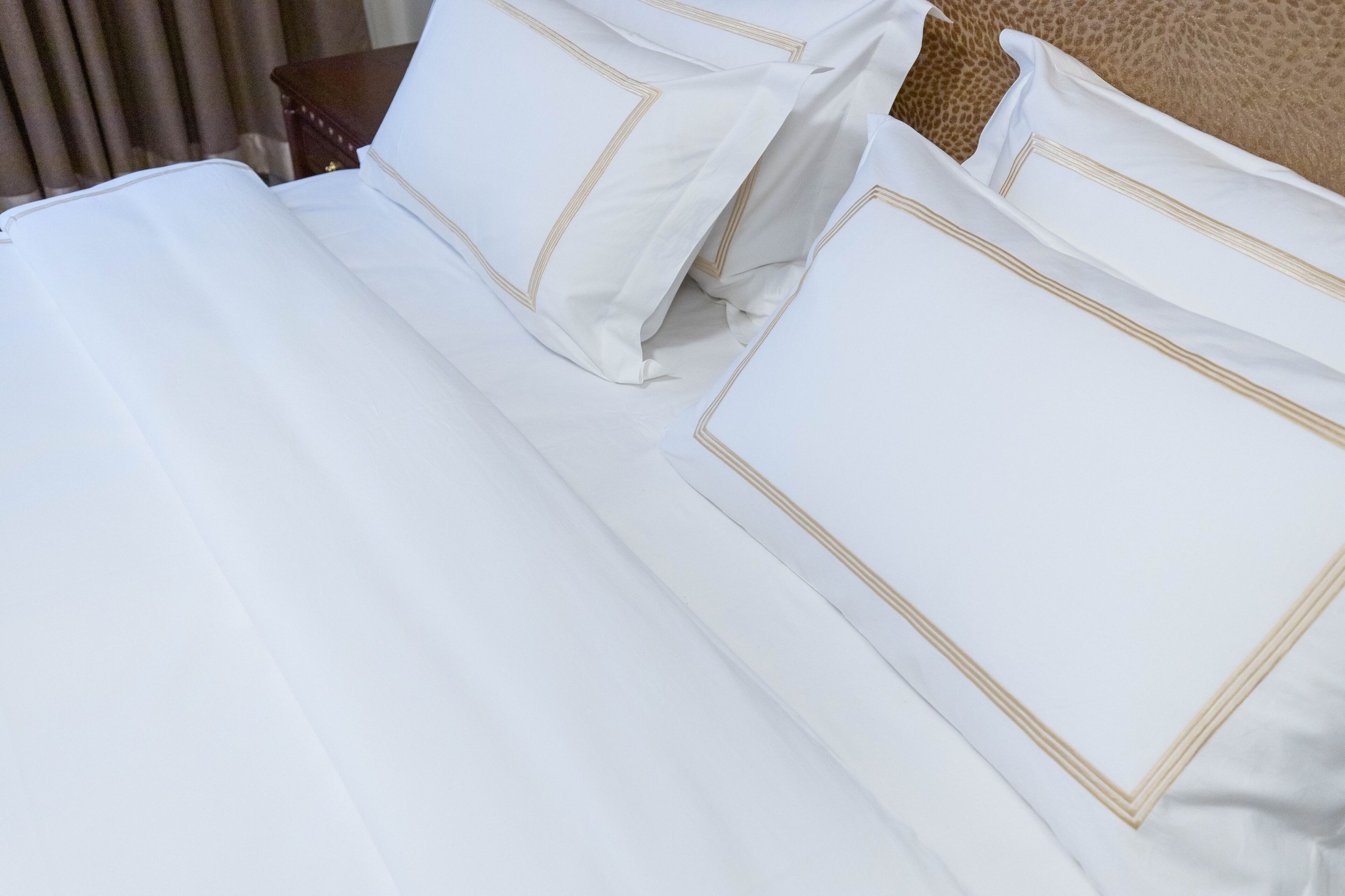 A neatly made bed with white linens and gold trim, a wooden nightstand, and a brown leather headboard.