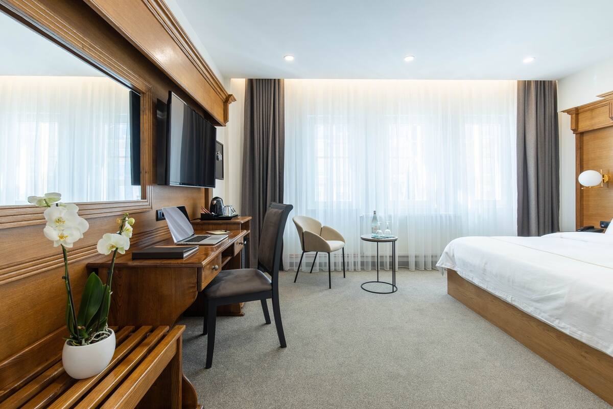 A hotel room with a bed, a desk with a laptop, a chair, a small table with a bottle, and a potted orchid.