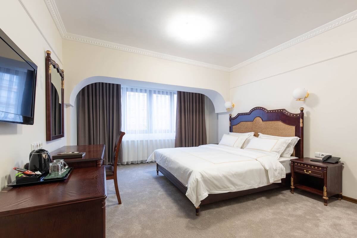 A hotel room with a large bed, a desk with a coffee maker, a TV, and a window with curtains.