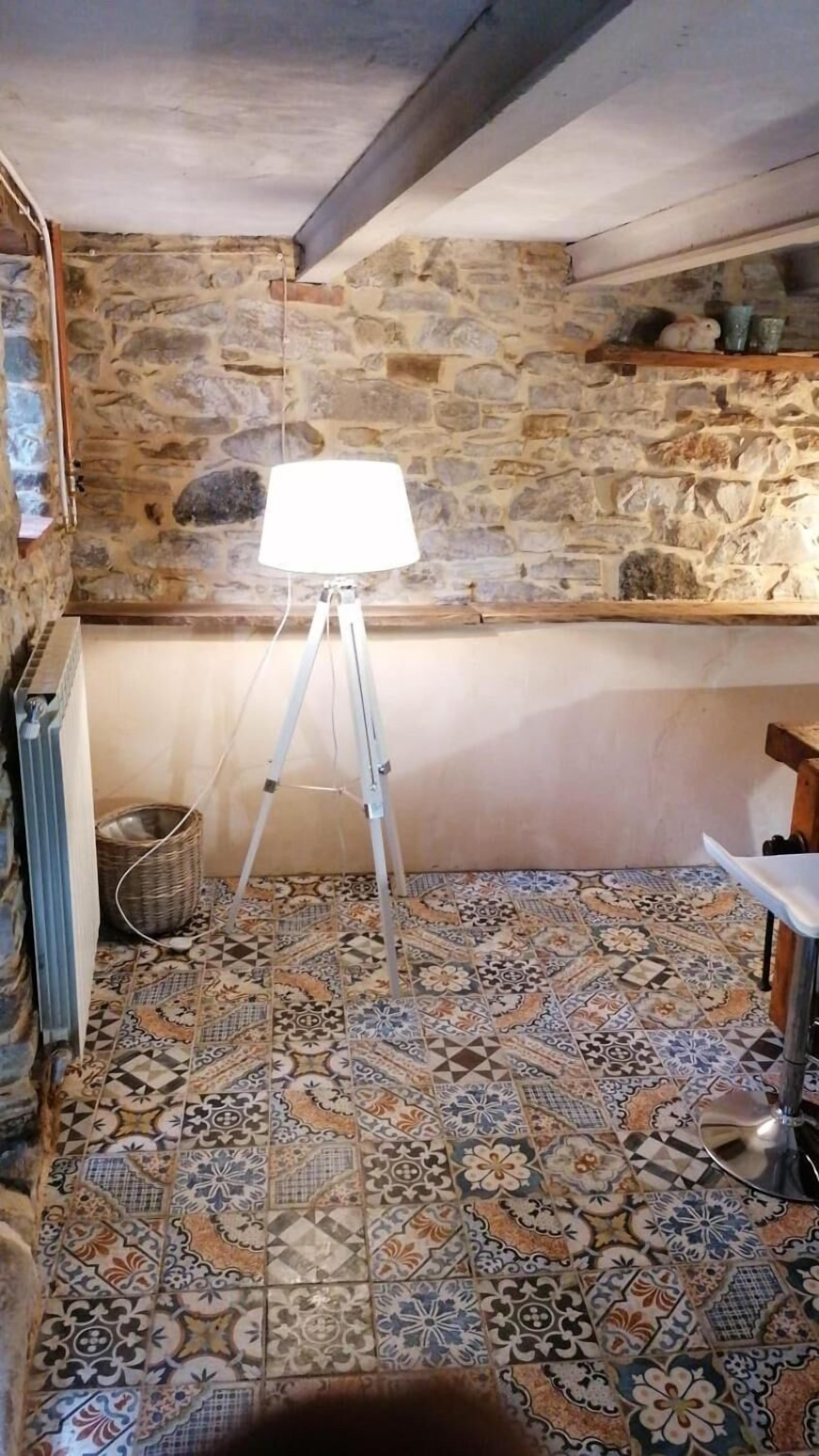 Una stanza con parete in pietra, un treppiede con lampada, un termosifone e un pavimento decorato.