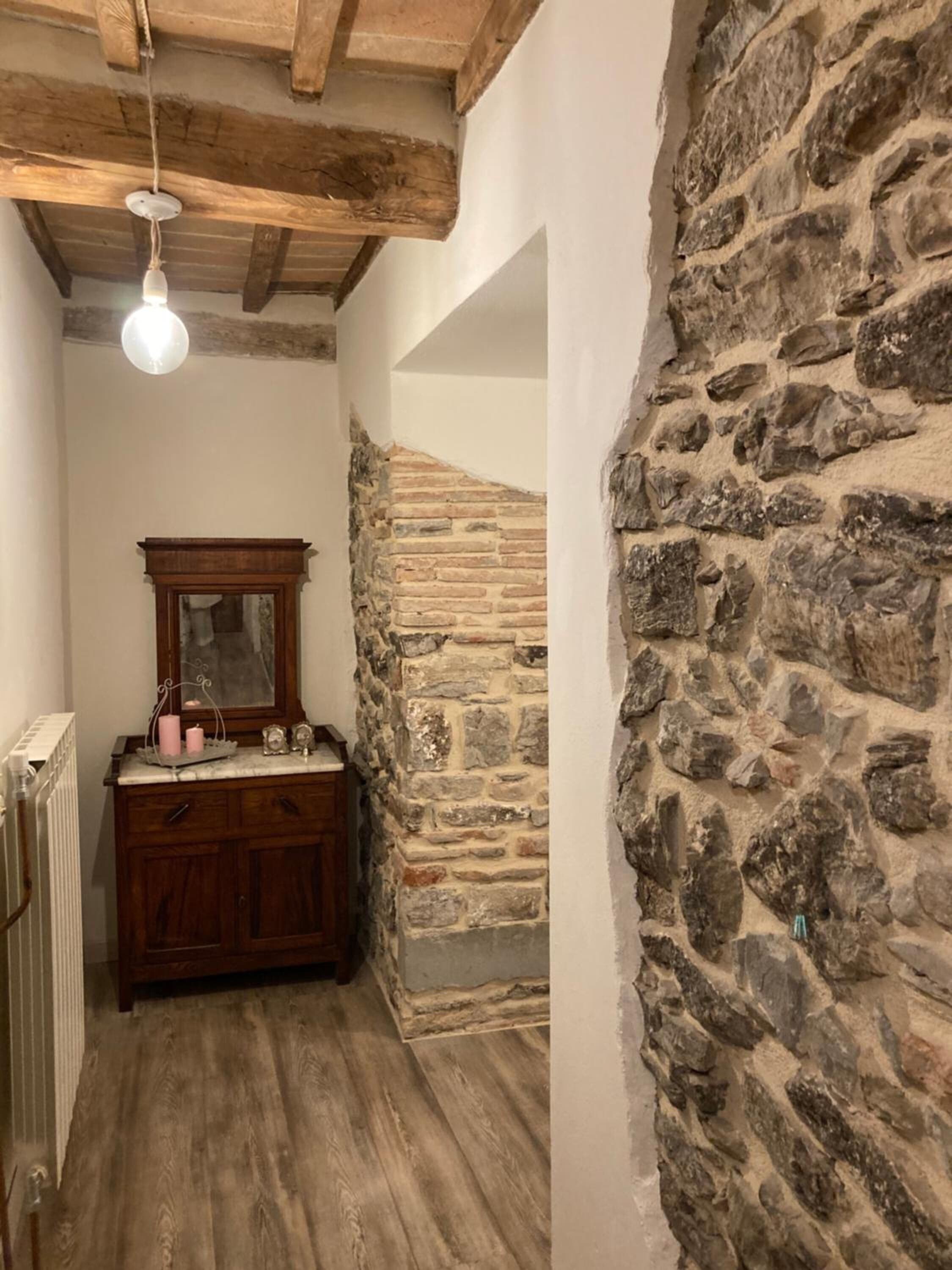 A narrow hallway with a stone wall, wooden beams, a wooden dresser, and a hanging light fixture.