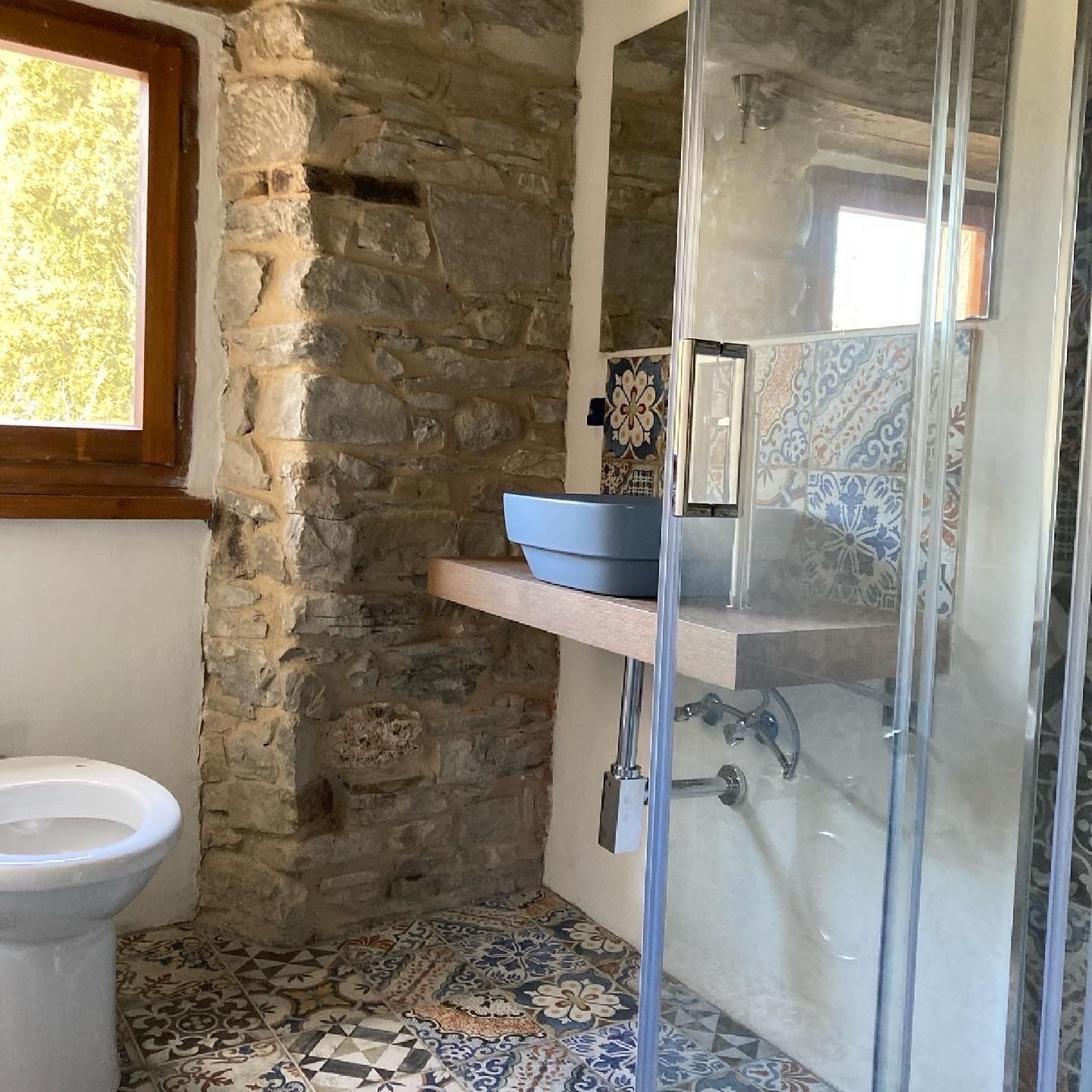 A bathroom with a stone wall, a tiled floor, a white sink, and a glass shower enclosure.