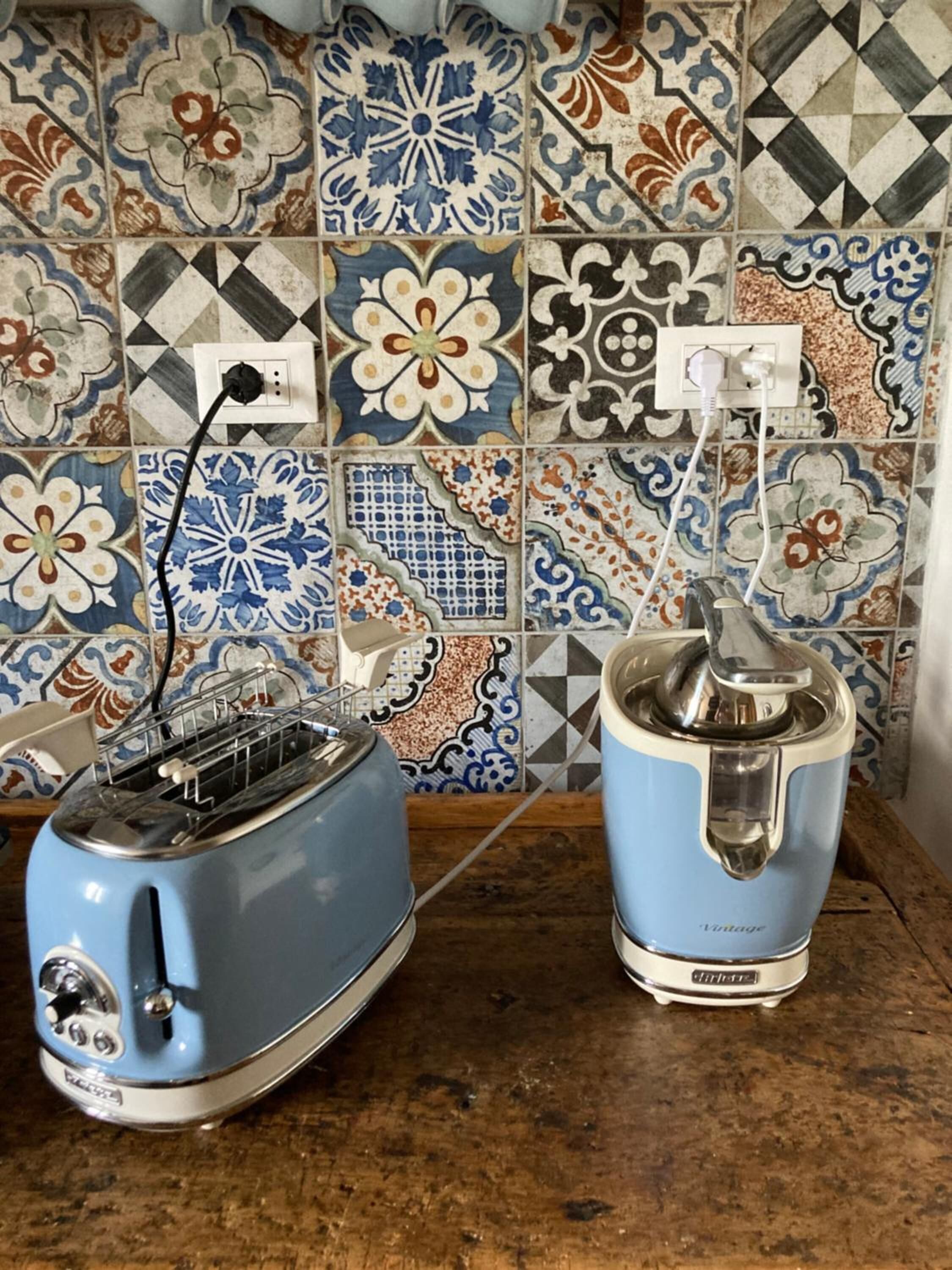 Une cuisine avec un grille-pain bleu et une cafetière ancienne posés sur un plan de travail en bois, le mur derrière étant recouvert de carreaux décorés de motifs floraux et géométriques.