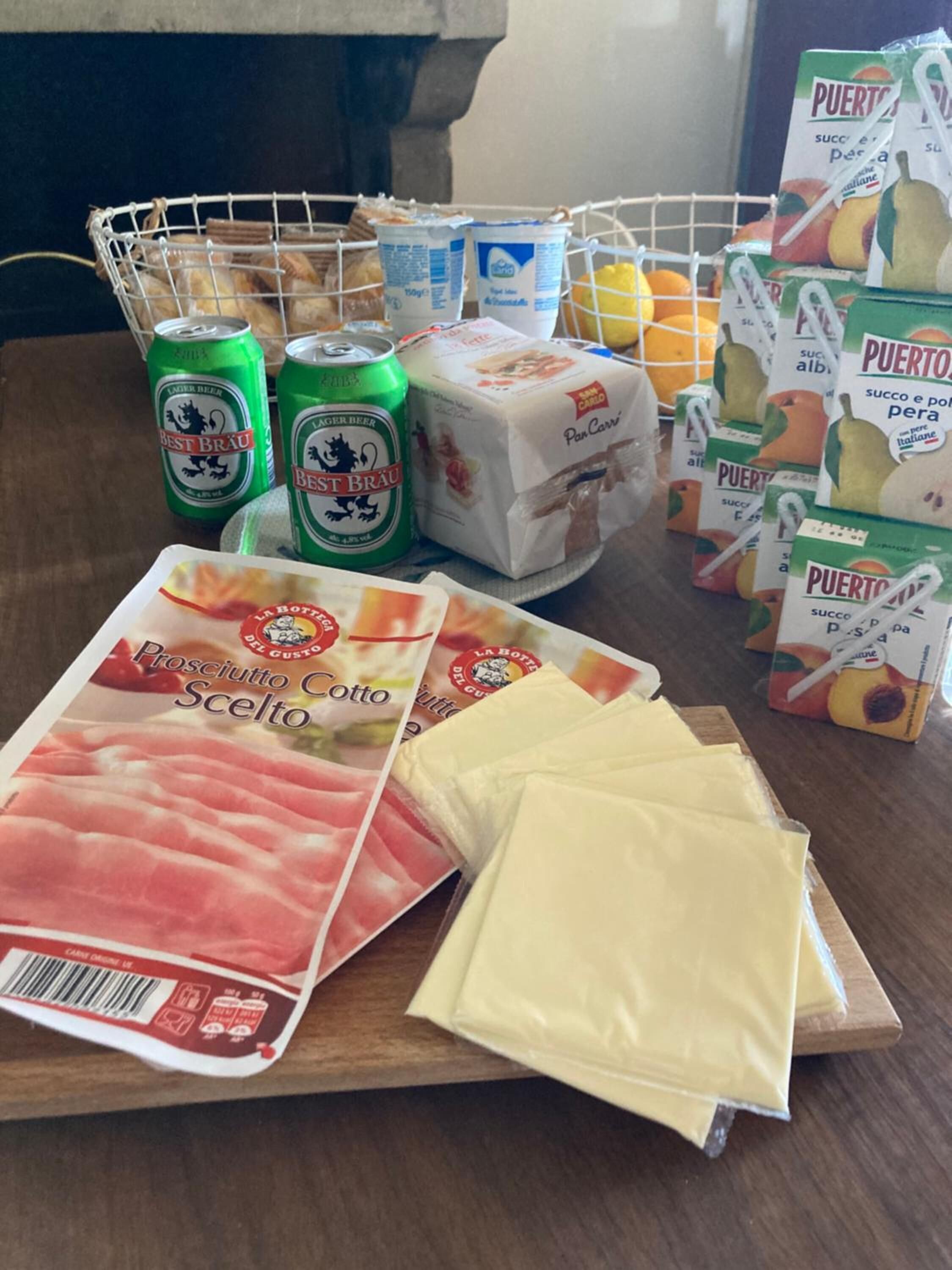Una cesta de compras con latas de cerveza, queso empaquetado y bolsas de fruta.