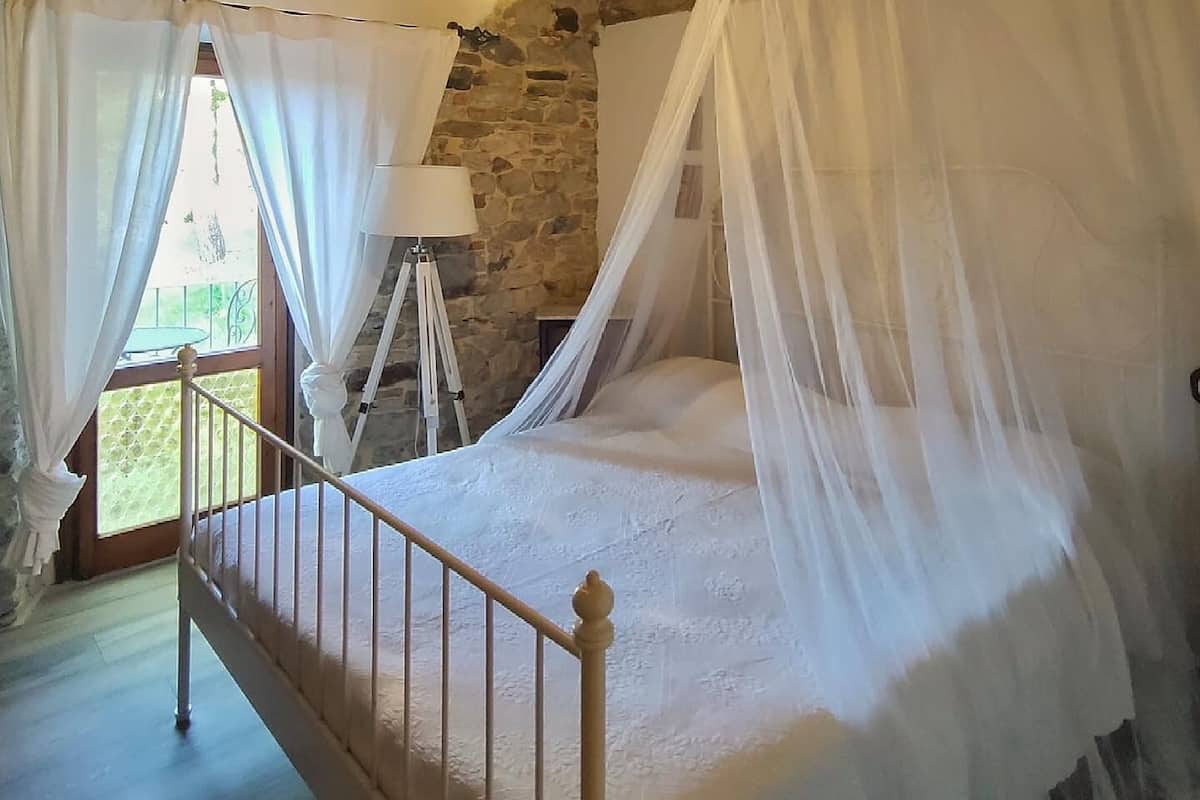 Un dormitorio con pared de piedra, cama con dosel, lámpara de pie y vistas a través de una ventana con cortinas.