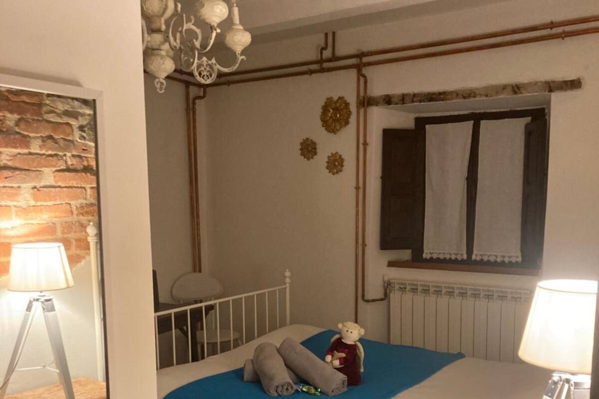 Un dormitorio con cama, mesita de noche, lámpara, cómoda, pared de ladrillo, candelabro y ventana con cortinas.