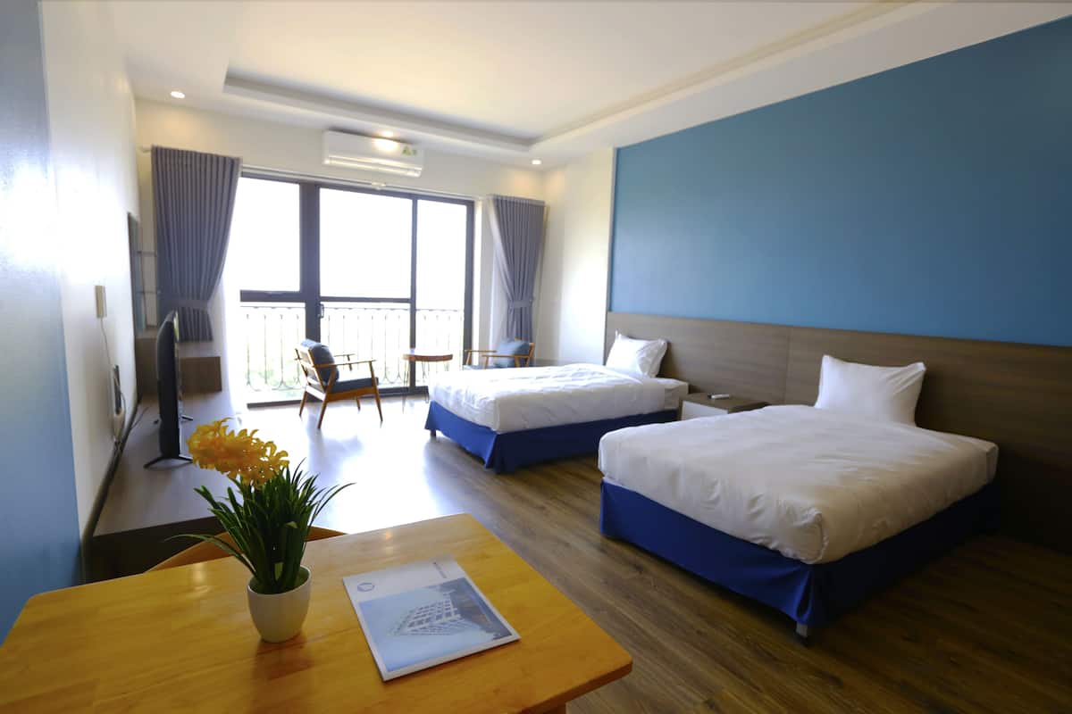 A hotel room with two beds, a wooden table with a plant, a balcony with a chair, and a large window.