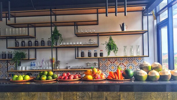 A kitchen counter with various fruits and vegetables, shelves with glassware and bottles, and a window with a view. - Minh Quan Building (Vinh Phuc)