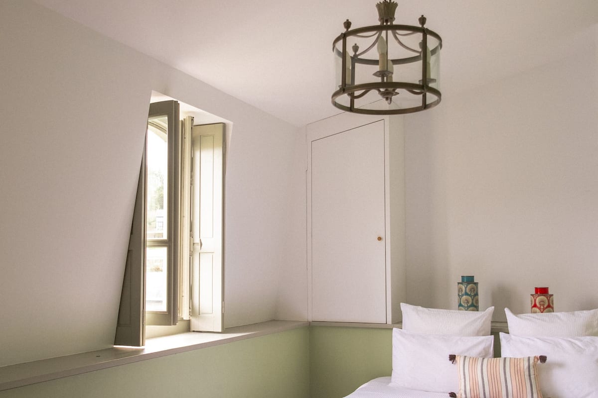 Un dormitorio con cama, almohadas, mesita de noche, ventana y puerta.