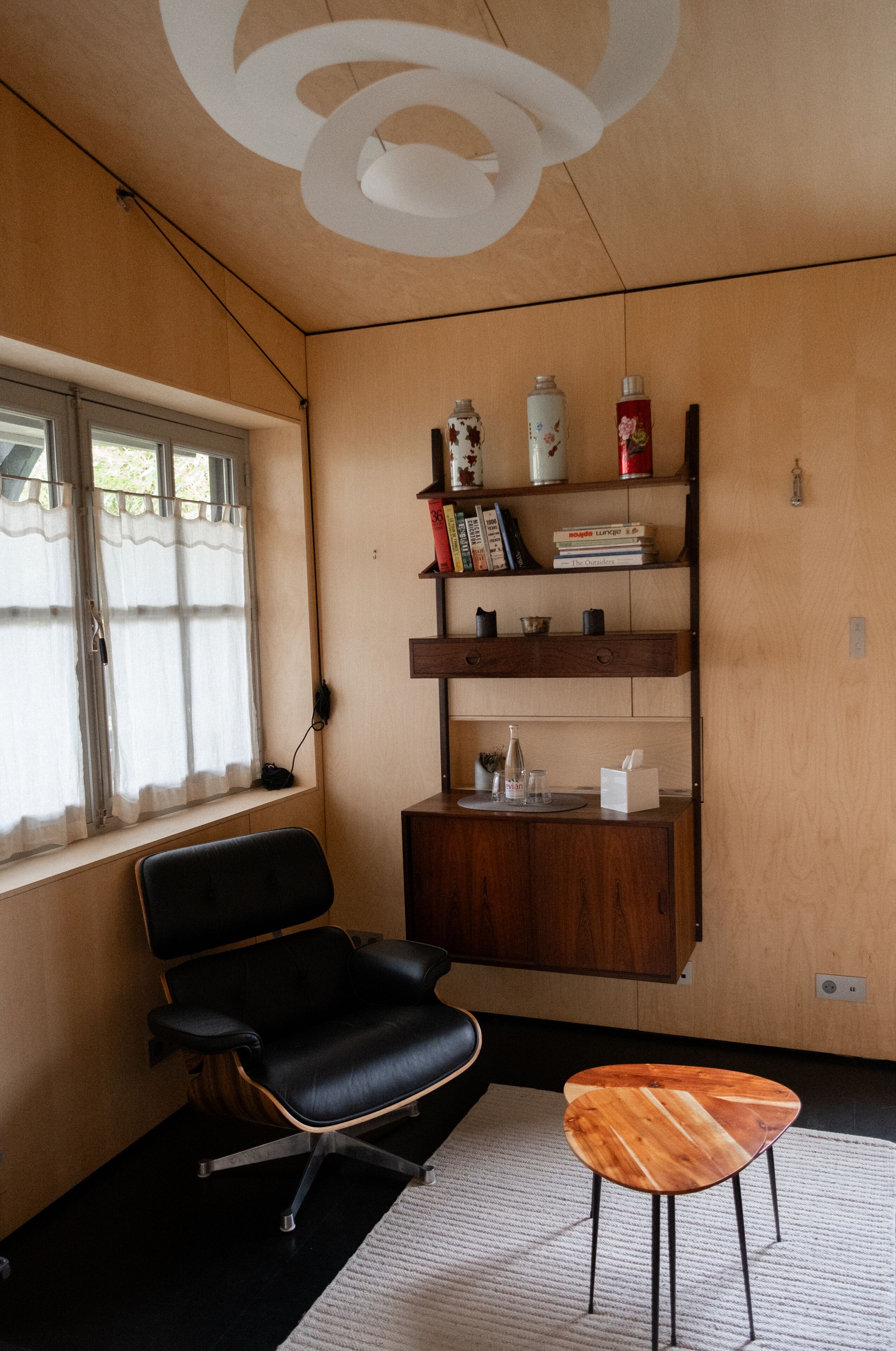 A room with a black leather chair, a wooden side table, a wooden bookshelf, and a window with a curtain.