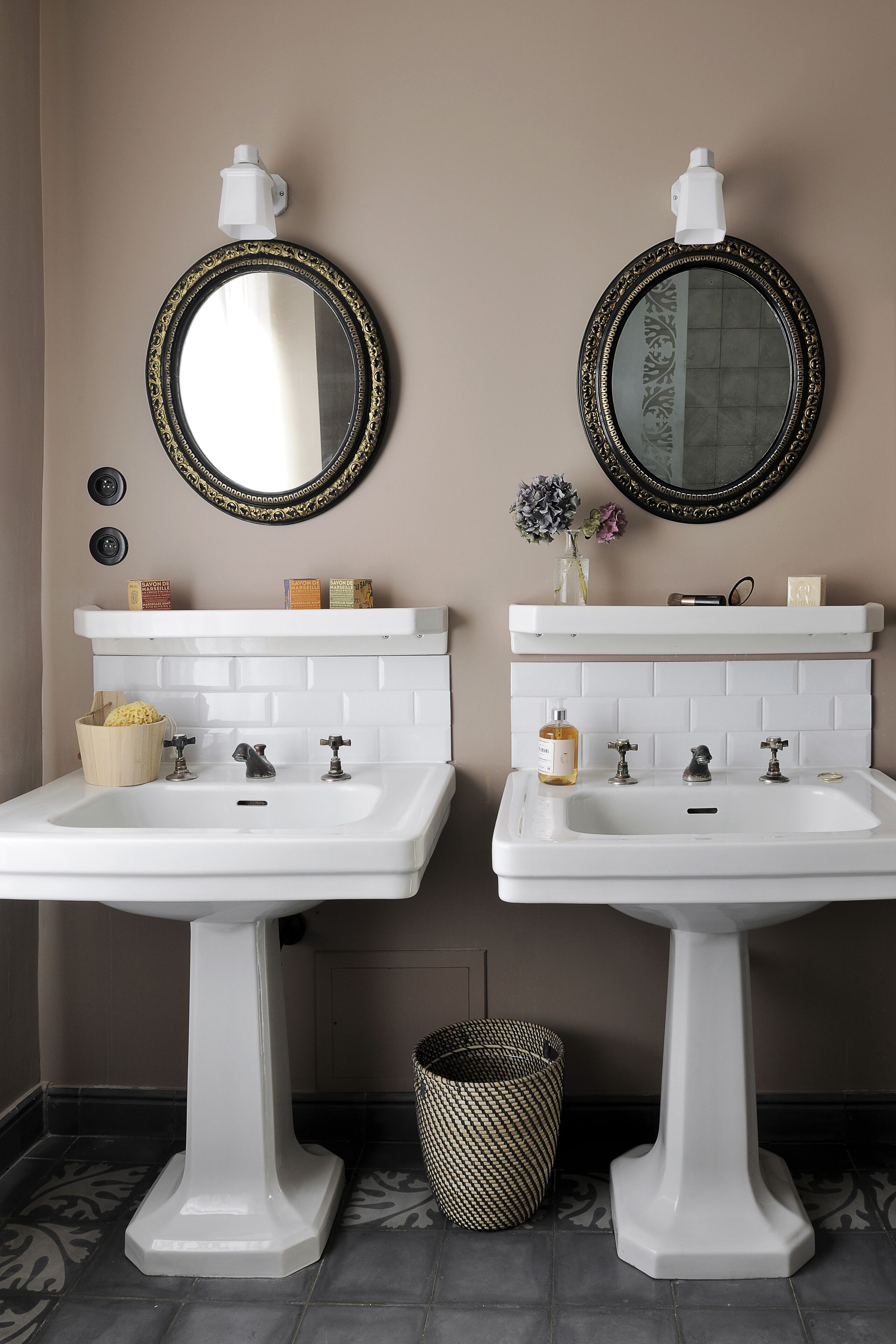 Two white pedestal sinks with gold-framed mirrors above them, a white shelf with toiletries, and a patterned floor.