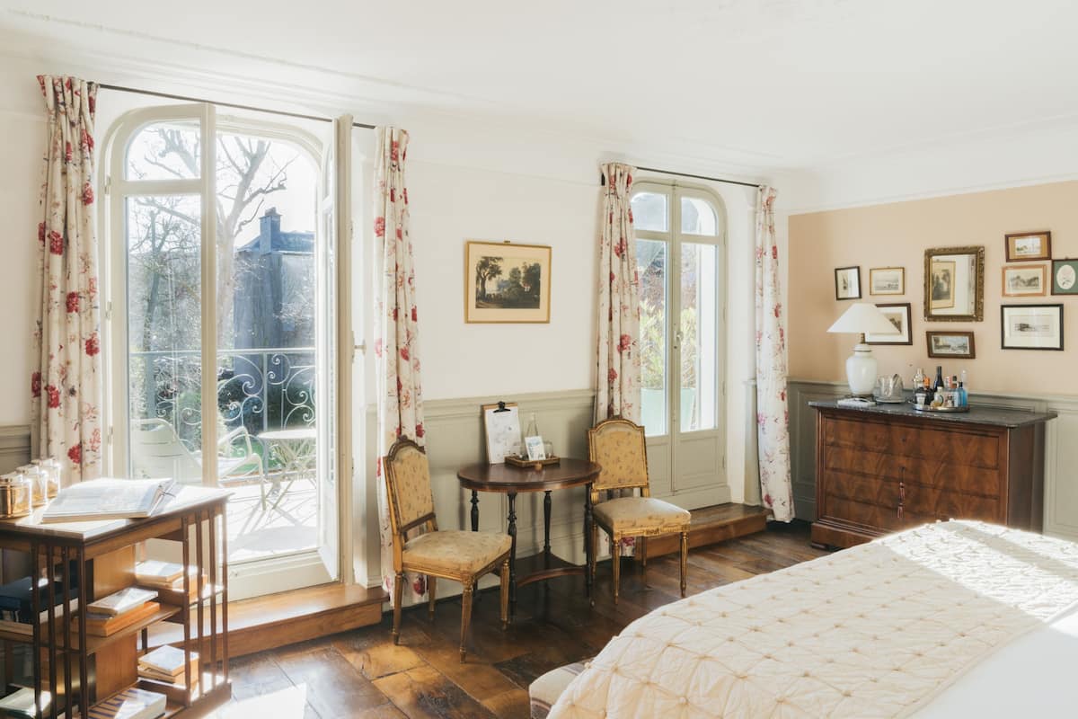 Una habitación con un ventanal grande, una cama, un escritorio con libros, dos sillas y un aparador con marcos de fotos.
