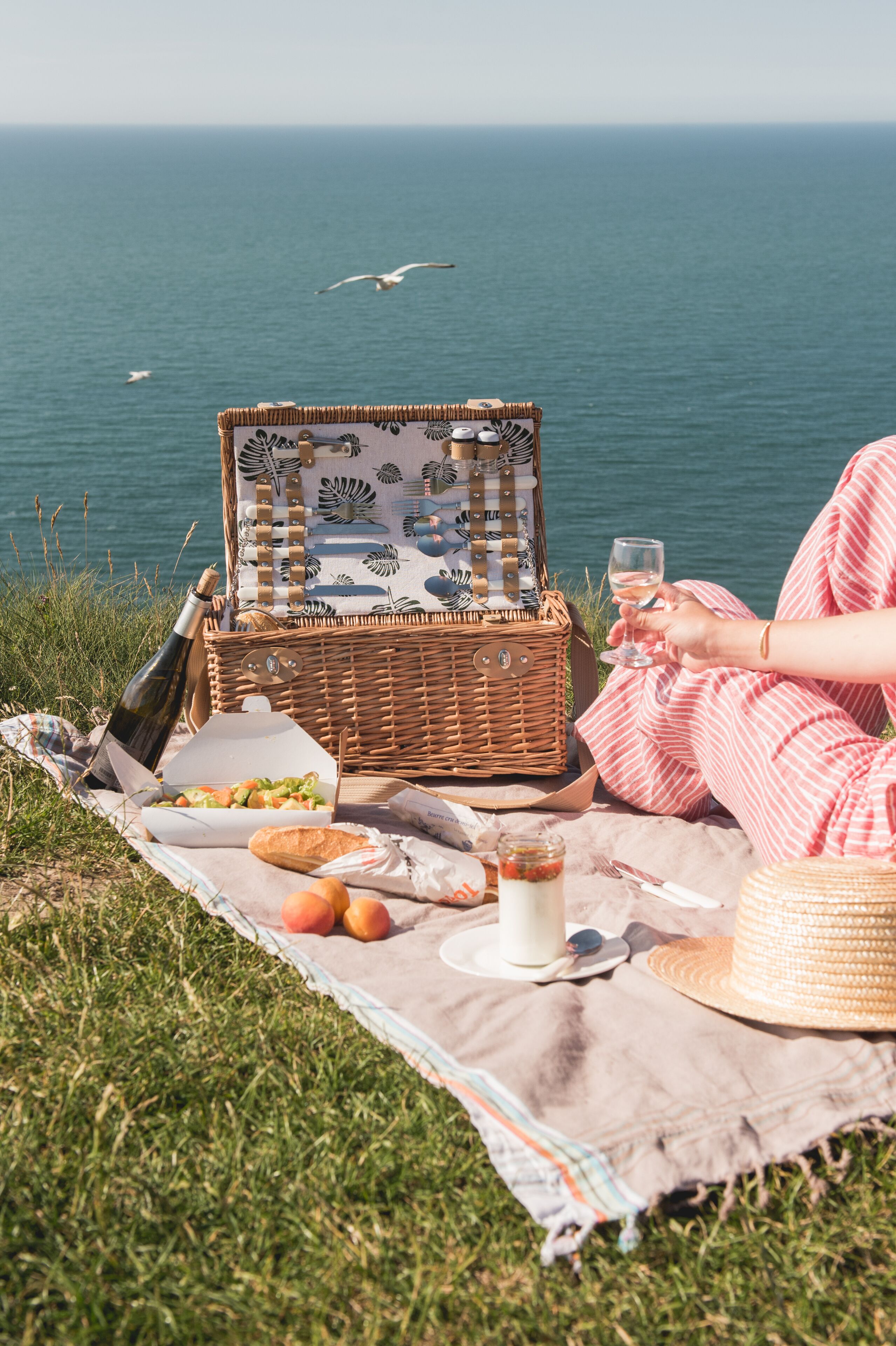 A picnic setup on grass with a wicker basket, food, and a glass of wine, overlooking the sea.
