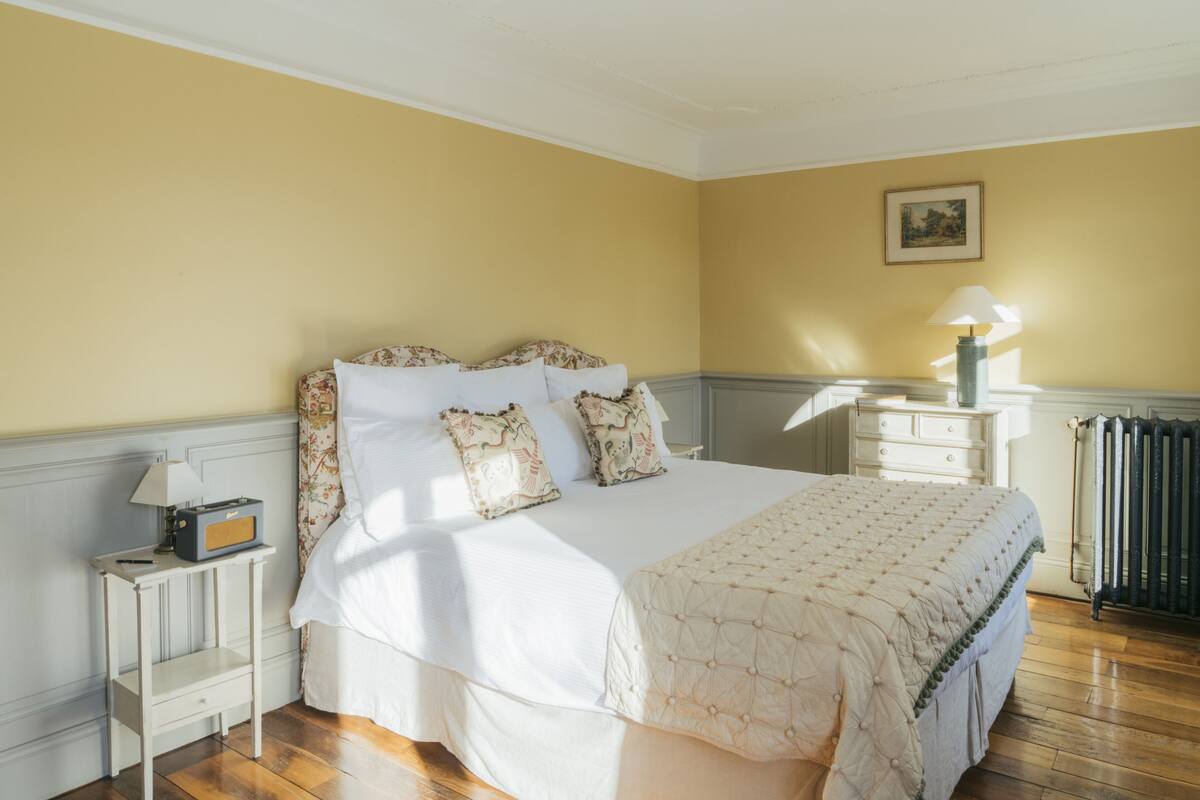 Una cama bien hecha con almohadas florales, una mesita de noche con una lámpara y un radiador en una habitación de paredes amarillas.
