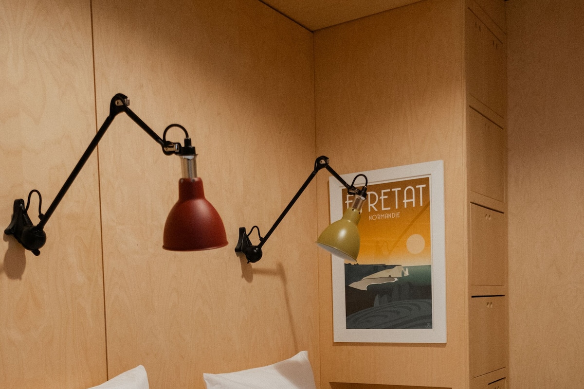 Una cama bien hecha con sábanas y almohadas blancas, una lámpara roja y verde fijada a la pared, y un cuadro en la pared.