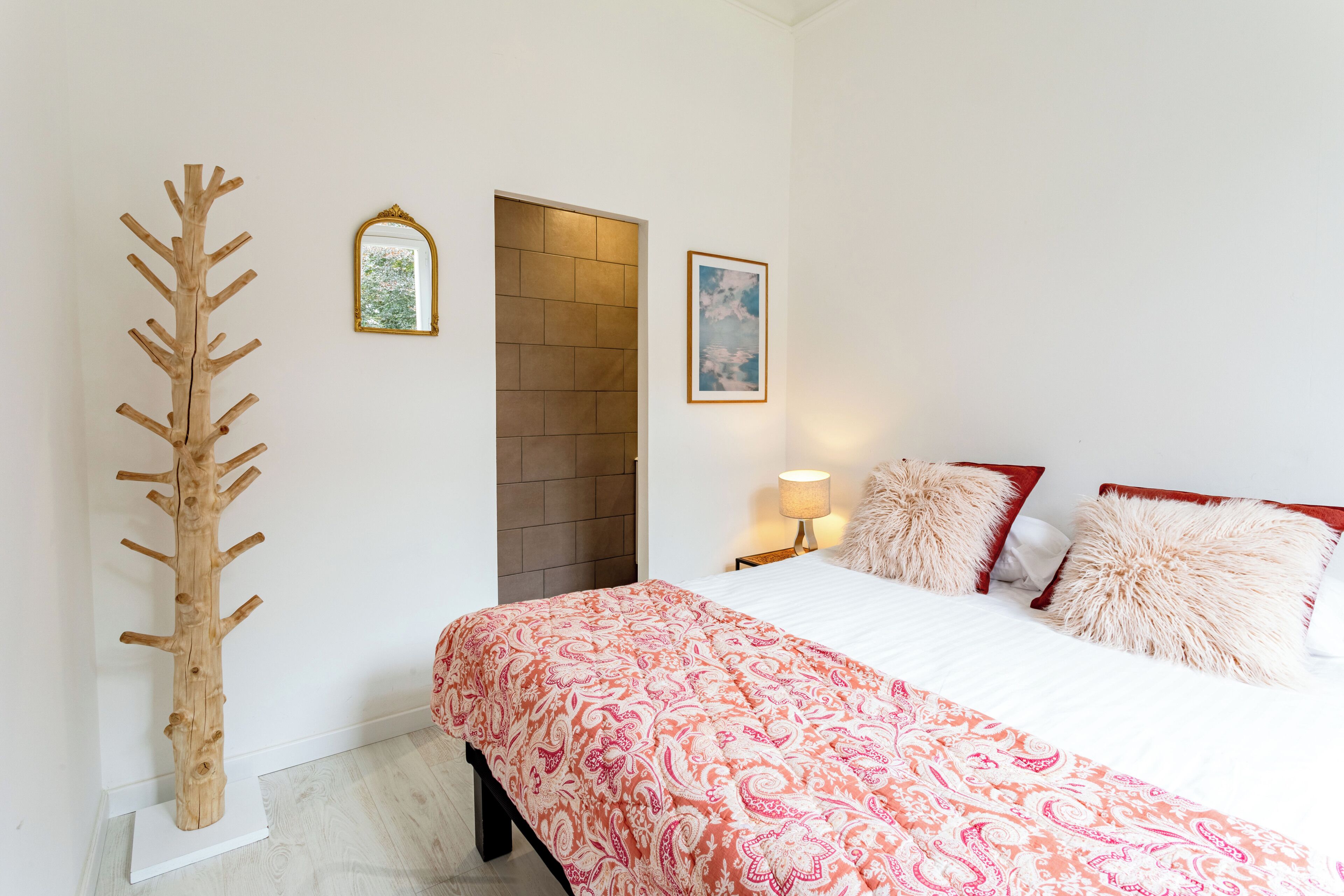 A bedroom with a bed, a wooden coat rack, a door, and two framed pictures on the wall.