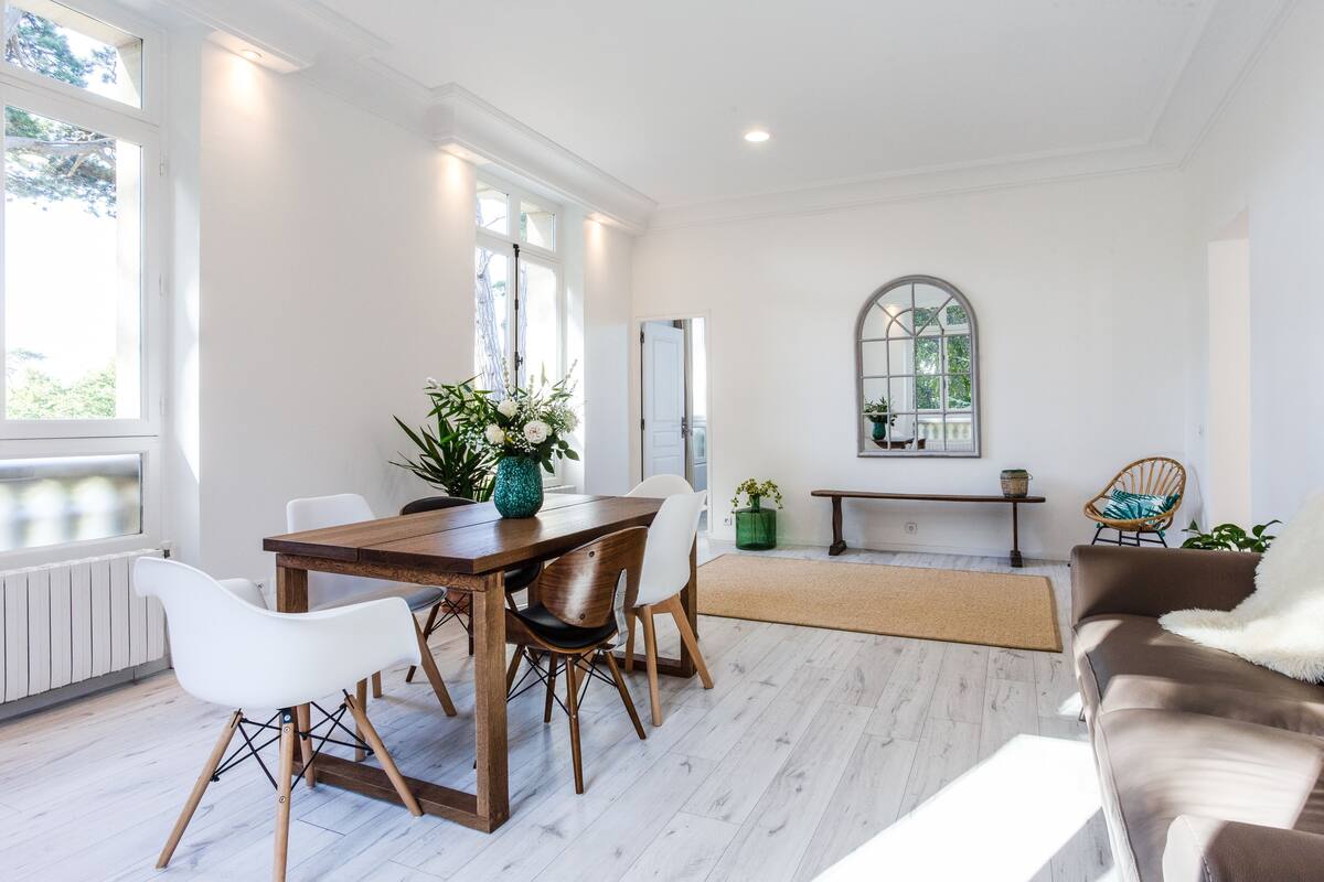 A spacious living room with a wooden dining table, white chairs, a brown sofa, and a large arched window.