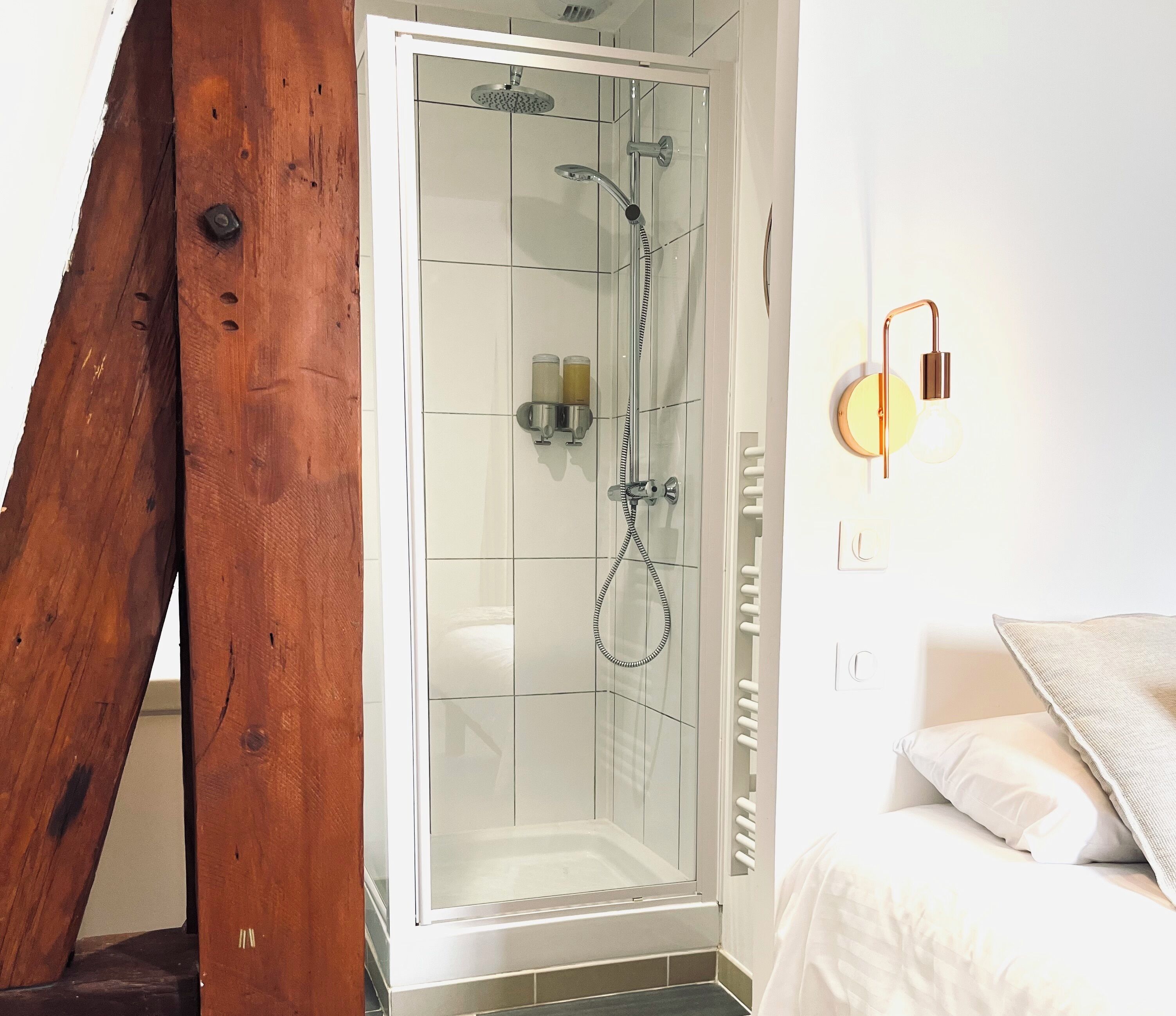 A modern bathroom with a glass shower enclosure, a wooden beam, and a bed with white bedding.