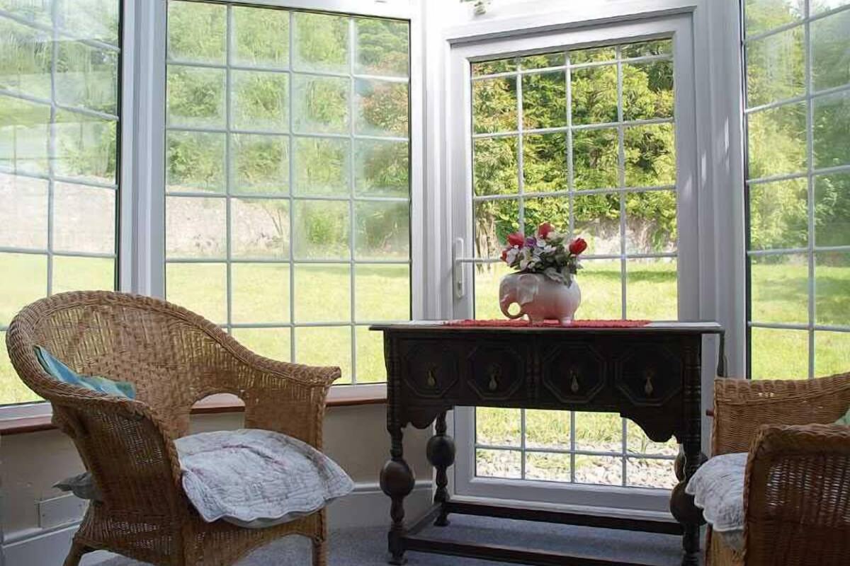 A room with large windows, wicker chairs, a small table with a vase of flowers, and a view of a green landscape.