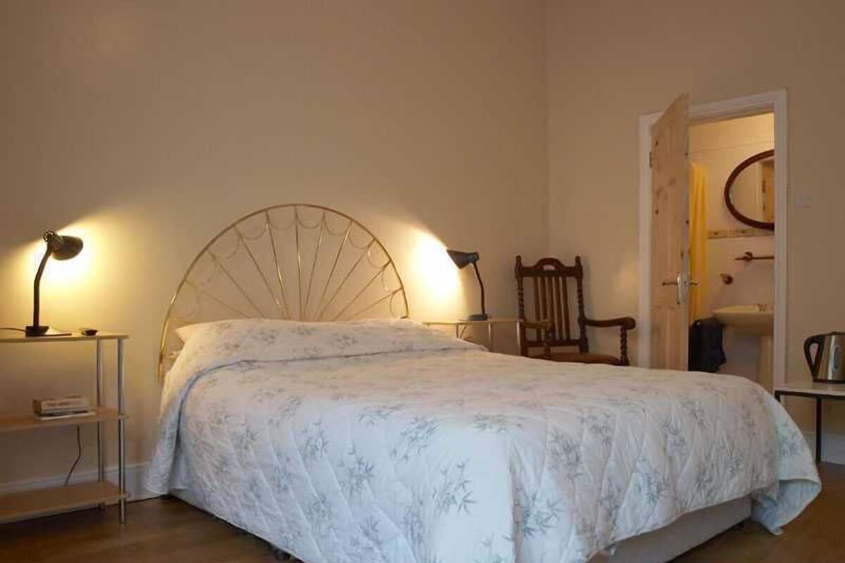 A bedroom with a bed, two bedside lamps, a wooden chair, and a door leading to a bathroom.