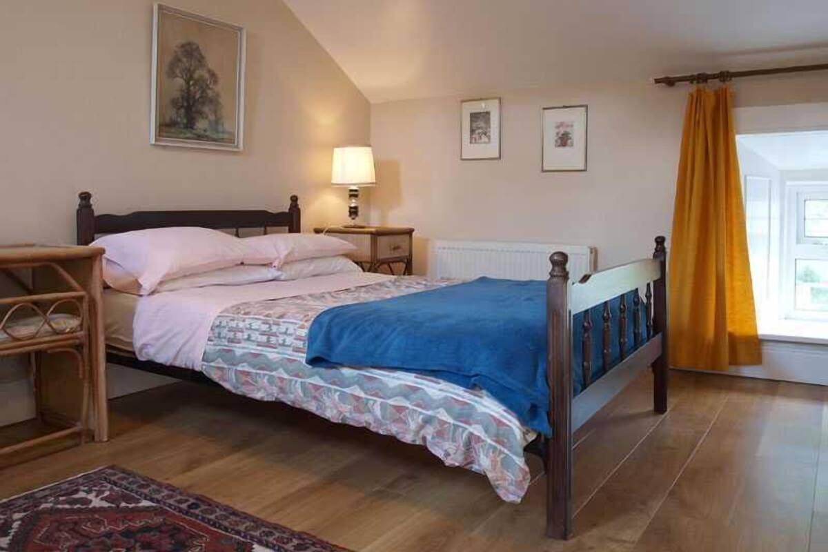 A bedroom with a bed, a nightstand, a lamp, a window with yellow curtains, and a painting on the wall.