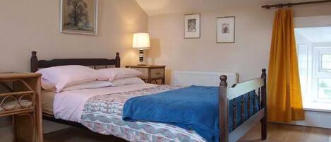 A bedroom with a bed, a nightstand, a lamp, a window with yellow curtains, and a painting on the wall.
