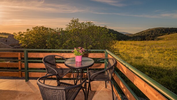 A balcony with wicker chairs and a table, overlooking a grassy landscape and hills. - Villa Windmill Sunset (Zakynthos)
