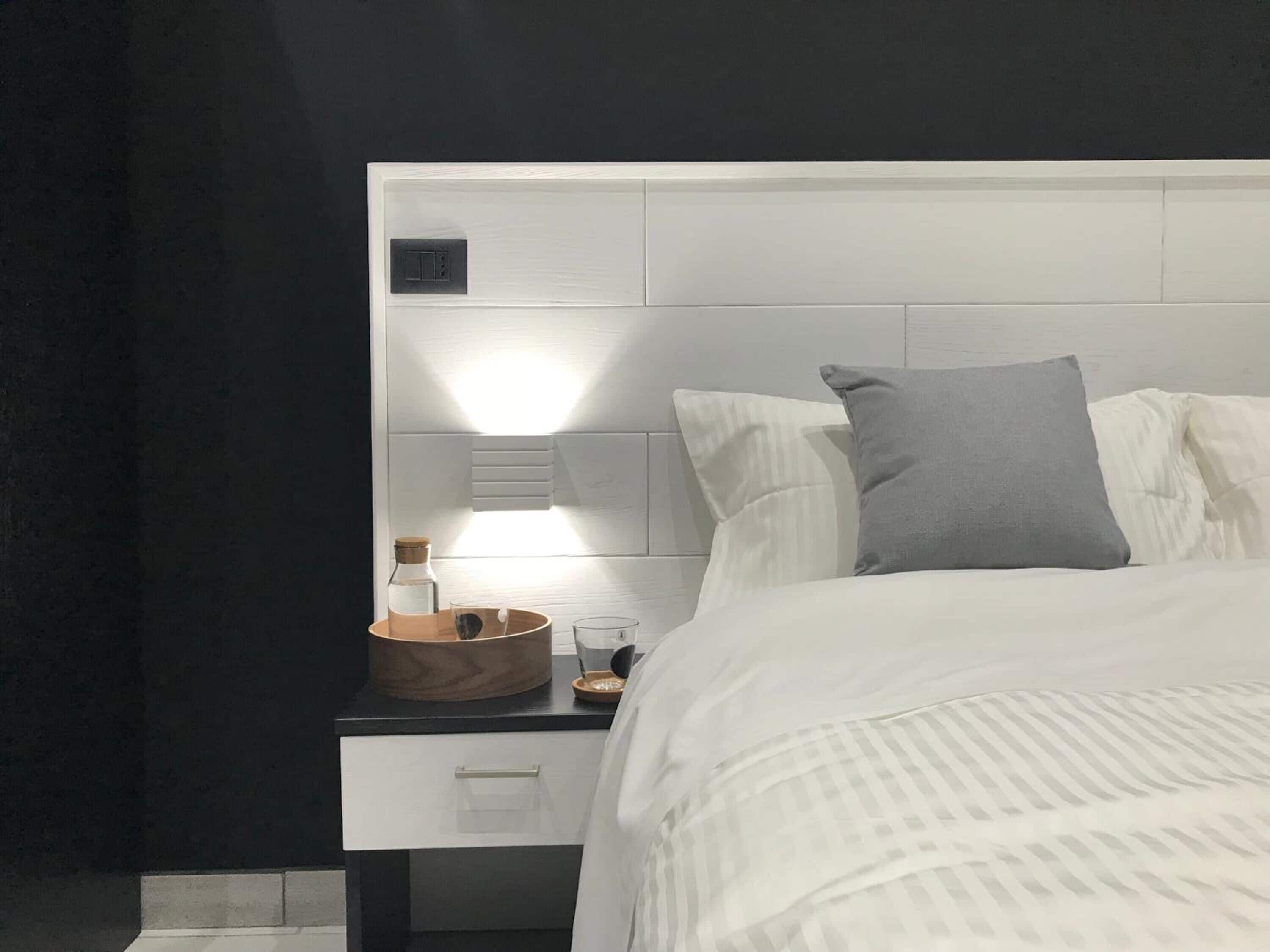Een netjes opgemaakt bed met wit beddengoed en een grijs kussen, een houten nachtkastje met een glas water en een klein kommetje, en een witte nachtkast met een lade en een lichtschakelaar.