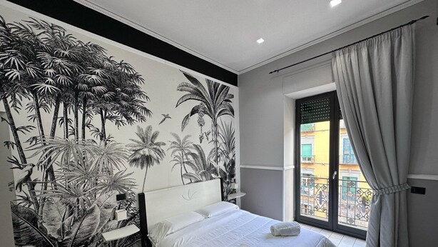 Un dormitorio con una cama grande, una mesita de noche, un mural en la pared con motivos de plantas tropicales y una ventana con cortinas.