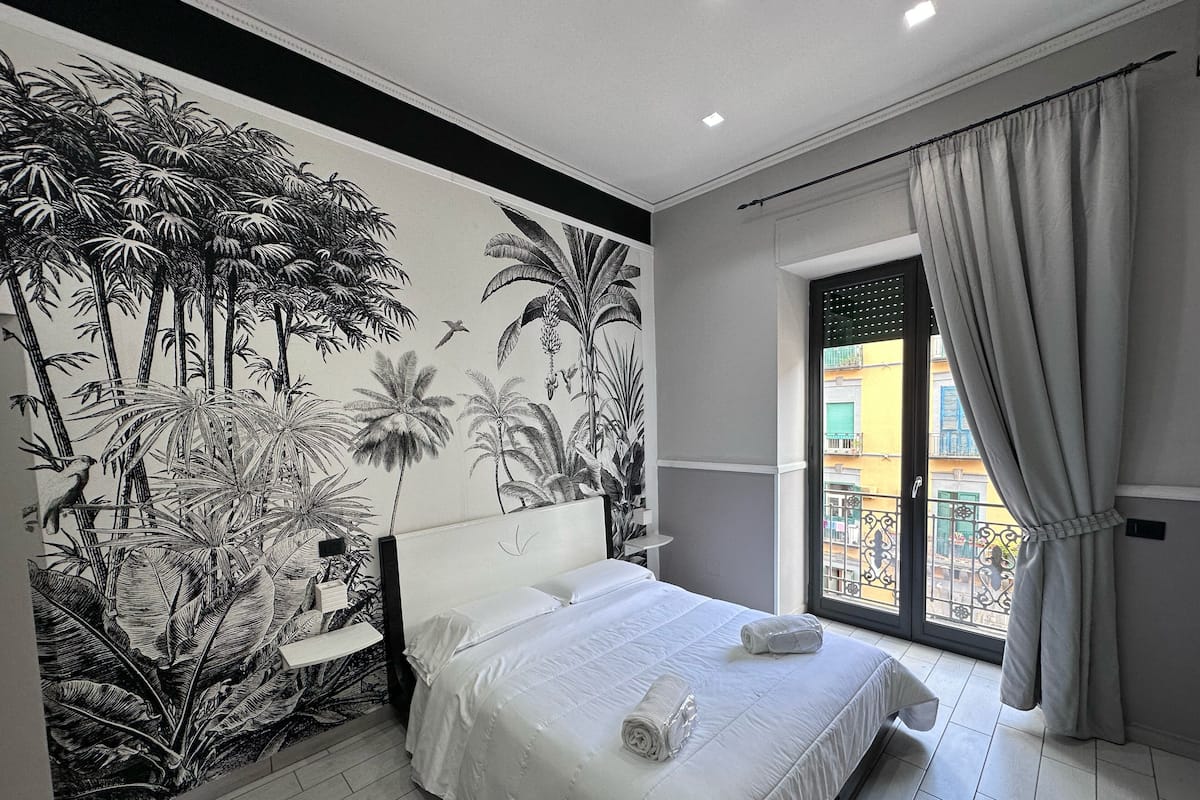 Una camera da letto con un letto grande, un comodino, un affresco floreale tropicale e una finestra con tende.