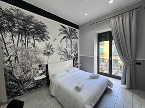 A bedroom with a large bed, a nightstand, a wall mural of tropical plants, and a window with curtains. - Parthenope Rooms (Naples)