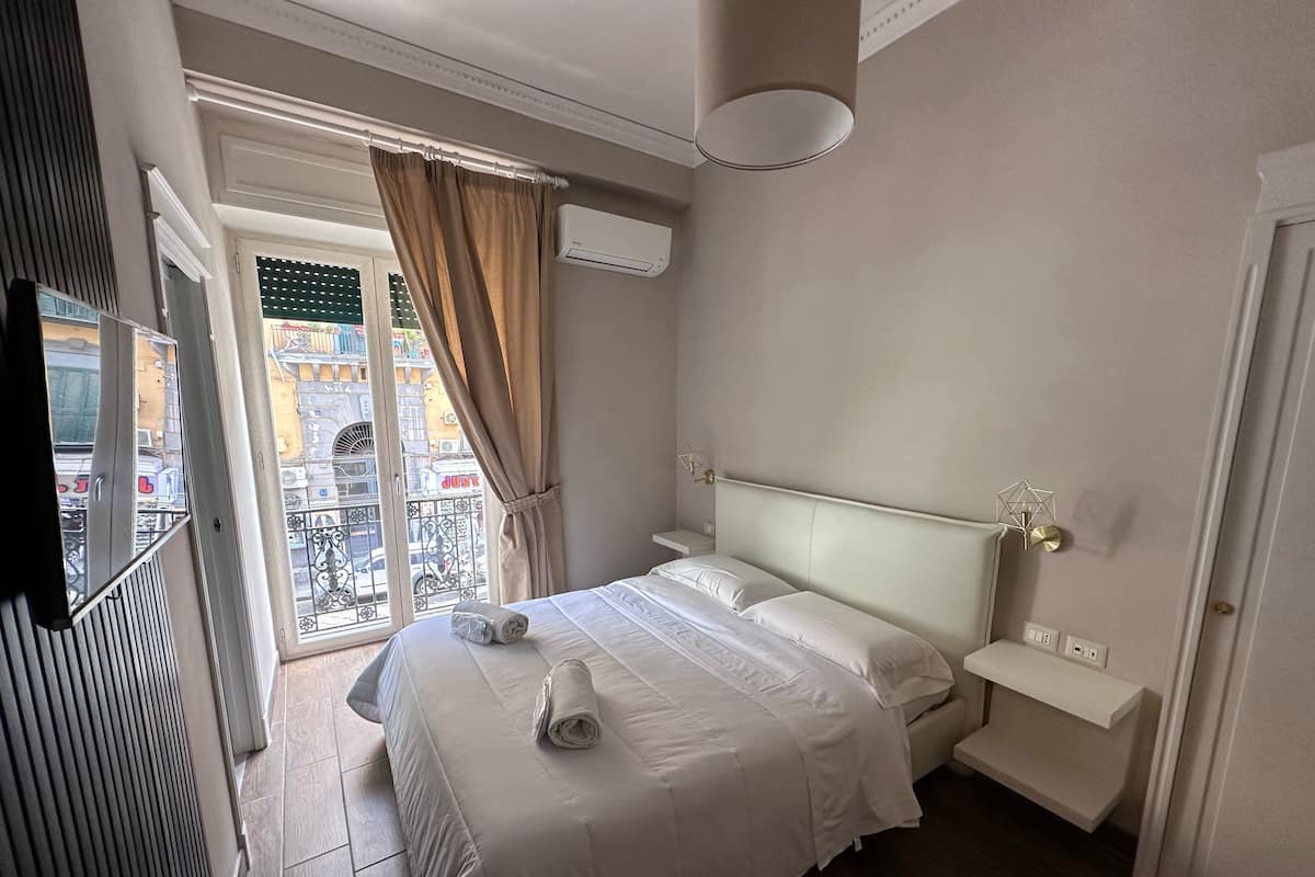 Una camera d'albergo con un letto, un comodino, una lampada a muro e una finestra con tende.