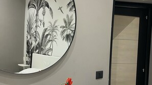 Un baño con tocador blanco, espejo redondo, un jarrón con una flor y un mural con motivos de hojas tropicales y pájaros.