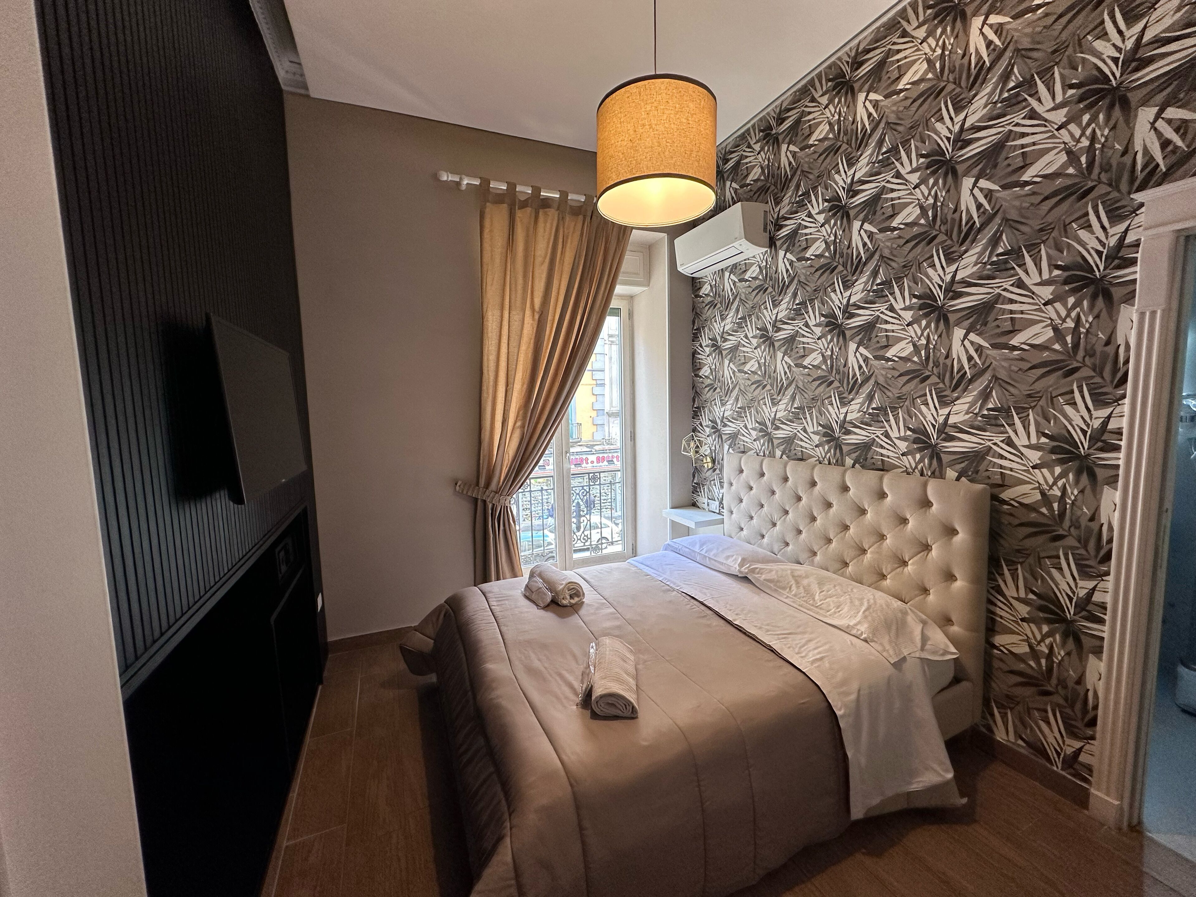 A bedroom with a tufted headboard, a flat-screen TV, a window with curtains, and a patterned wall.