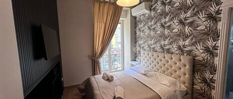 A bedroom with a tufted headboard, a flat-screen TV, a window with curtains, and a patterned wall.
