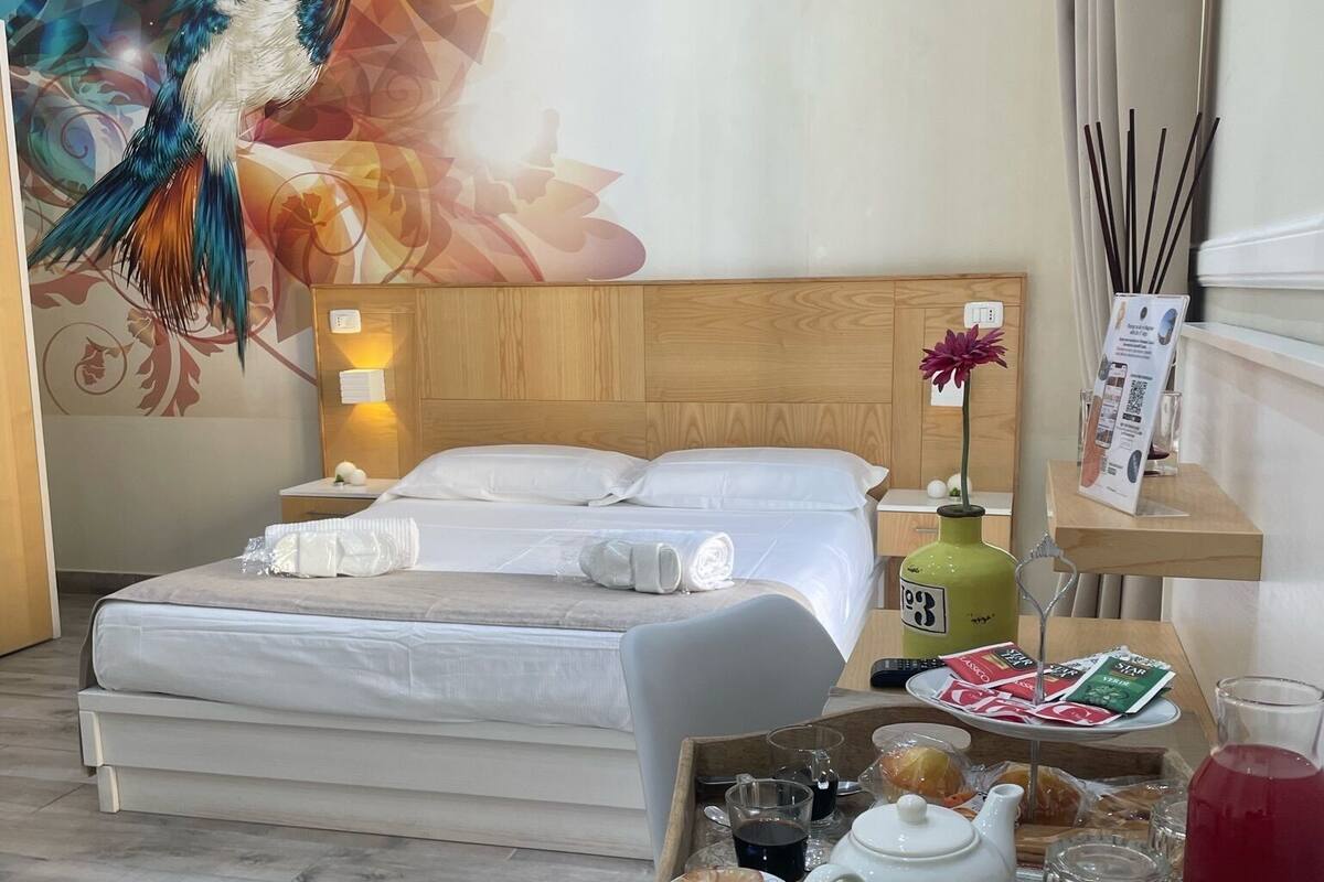 Una camera d'albergo con una testiera in legno, un letto con lenzuola bianche, un vassoio con articoli per la colazione, un vaso verde con un fiore e un affresco raffigurante un uccello.
