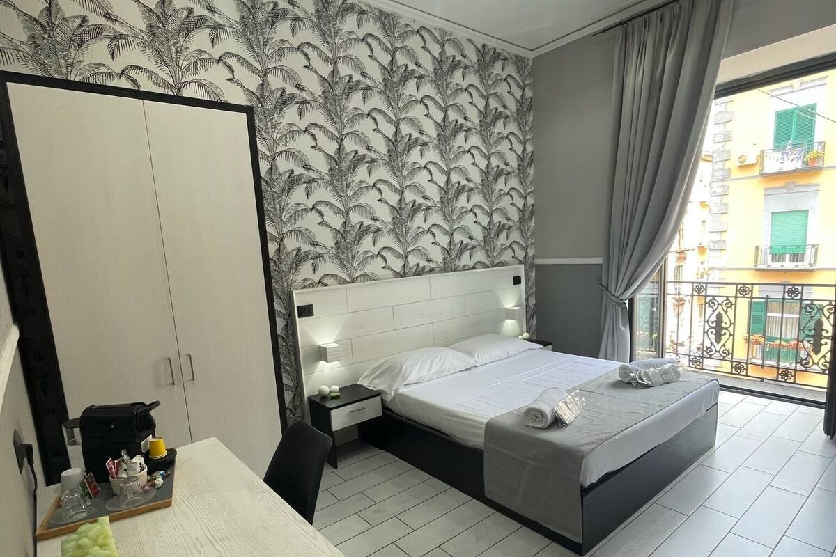Camera d'albergo moderna con un letto grande, una scrivania con specchio, un armadio e vista sulla città da un balcone.