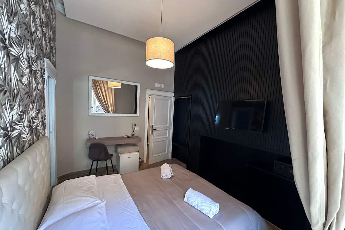 Una camera d'albergo con un letto, una scrivania, una sedia, uno specchio, una televisione e una finestra con tende.