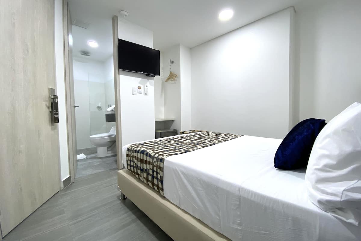 A modern hotel room with a bed, a flat-screen TV mounted on the wall, a bedside table, and a bathroom visible through an open door.