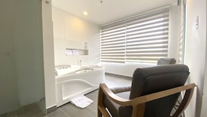 Ein modernes Badezimmer mit einer Badewanne, einem Waschbecken und einem Waschtisch. Ein Handtuch liegt auf dem Boden und ein Stuhl steht in der Nähe des Fensters.