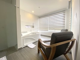 Un baño moderno con bañera, lavamanos y tocador. Hay una toalla en el suelo y una silla cerca de la ventana.