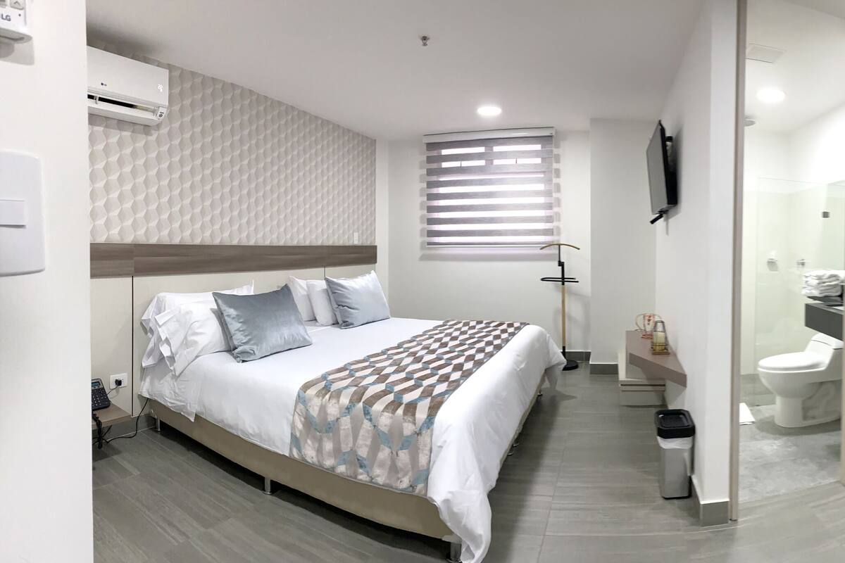 A modern hotel room with a bed, a black leather chair, a small table, and a bathroom visible through a glass partition.