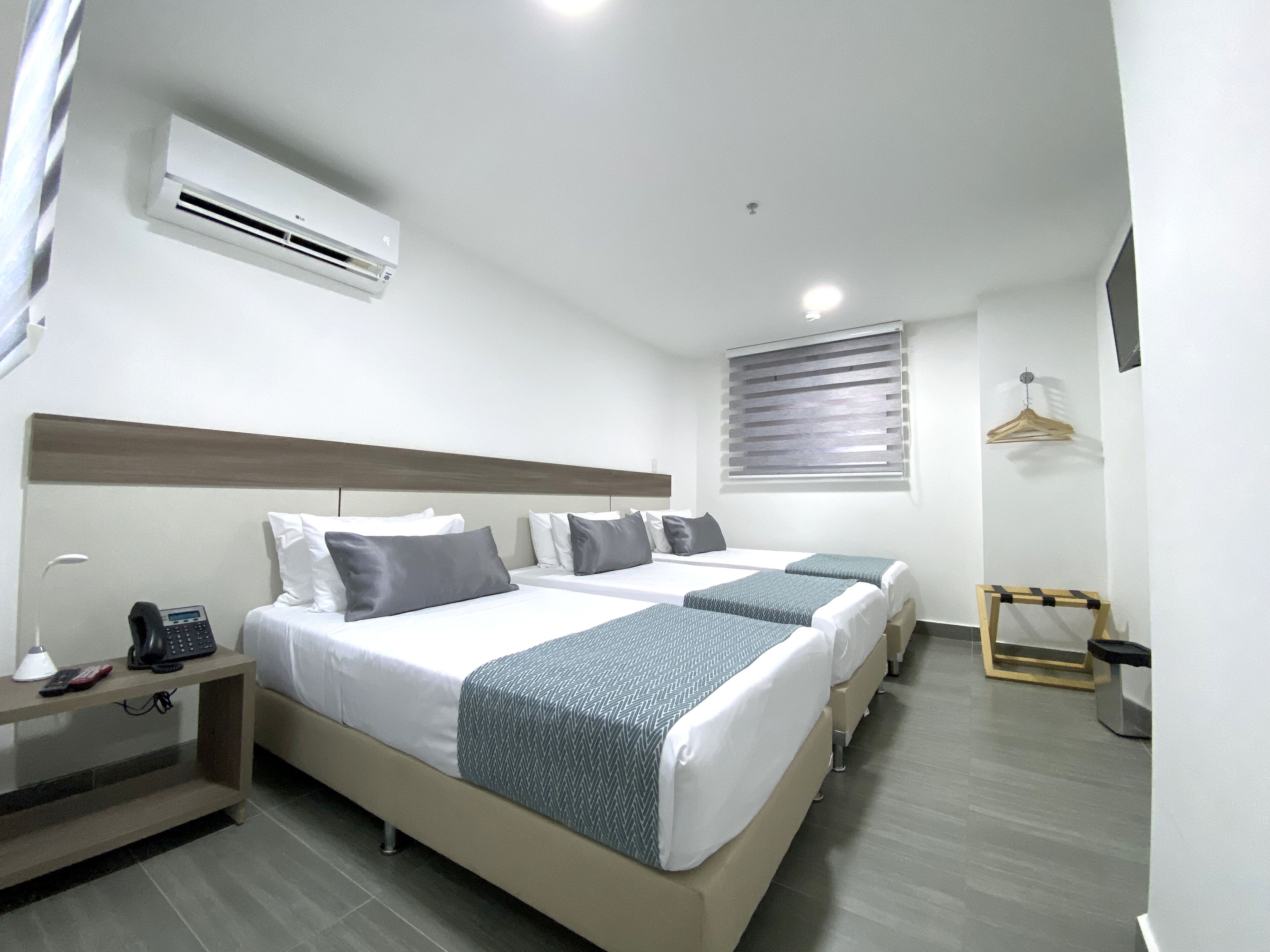 Une chambre d’hôtel avec deux lits, une table de chevet avec un téléphone et une lampe, un luminaire suspendu, une fenêtre avec des stores et un climatiseur fixé au mur.