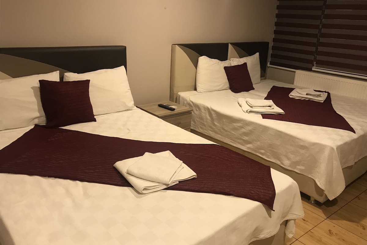 A hotel room with two beds, each with white linens and maroon pillows. There are folded towels on each bed, and a remote control is visible on the nightstand.
