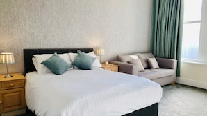 A hotel room with a large bed, a bedside table with a lamp, a small table with a phone, a sofa, and a window with curtains. - The Clarendon Hotel (Morecambe)