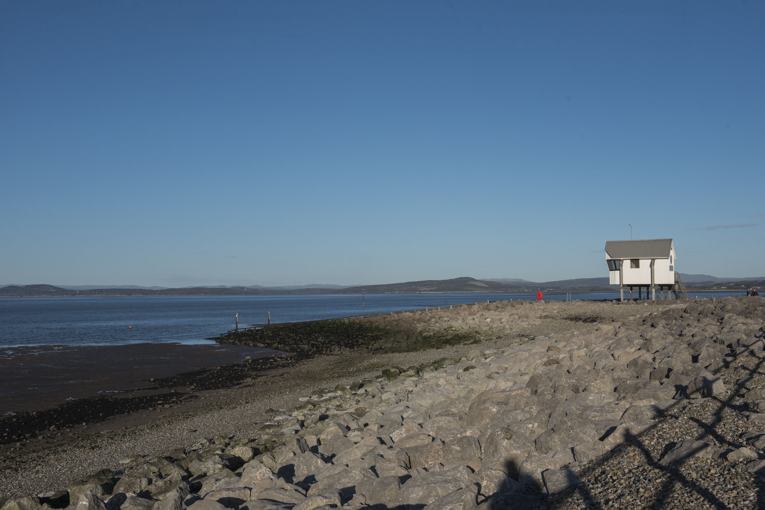 A rocky beach with a white building on stilts, overlooking a calm body of water.