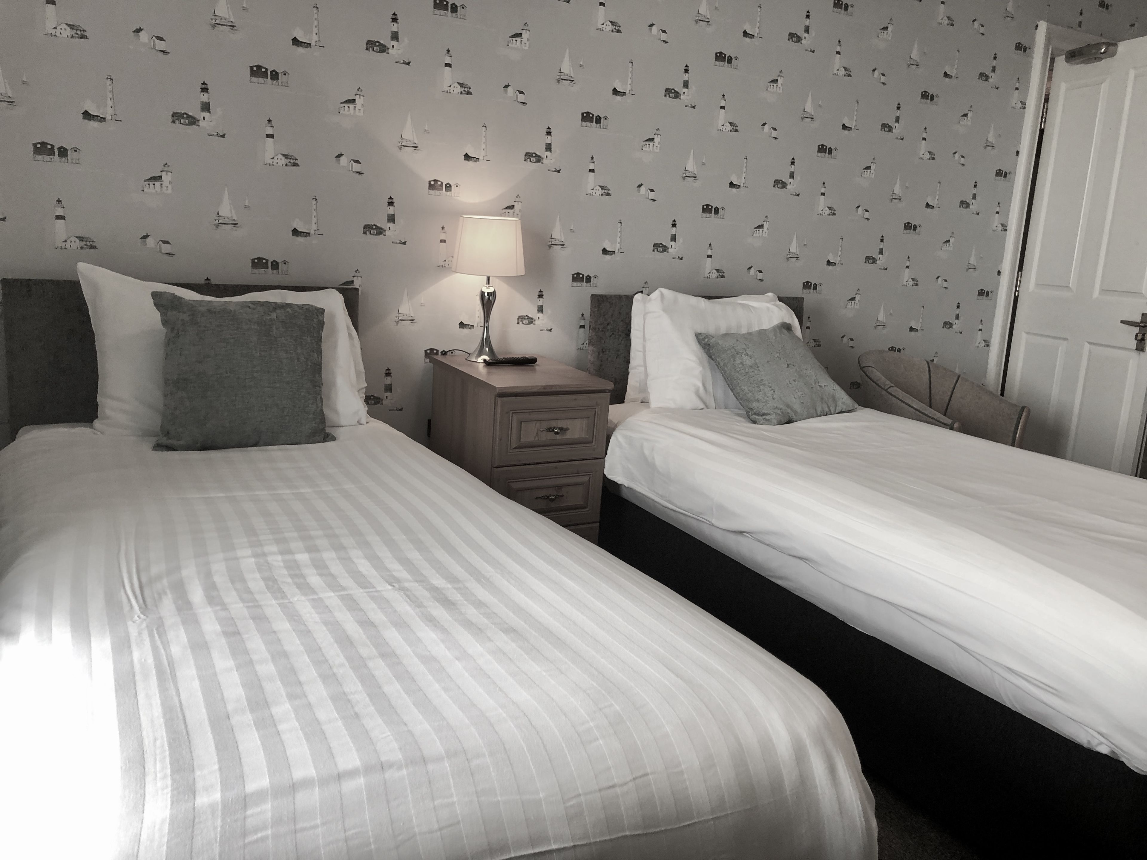 Una habitación con dos camas, una mesita de noche con lámpara y un papel tapiz de motivos náuticos.