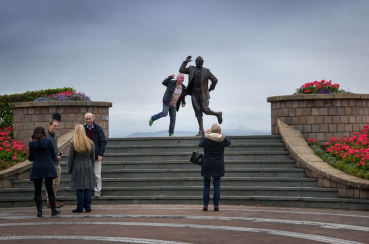 Una estatua de un hombre con traje y sombrero sobre una serie de escalones, con macizos de flores a ambos lados.