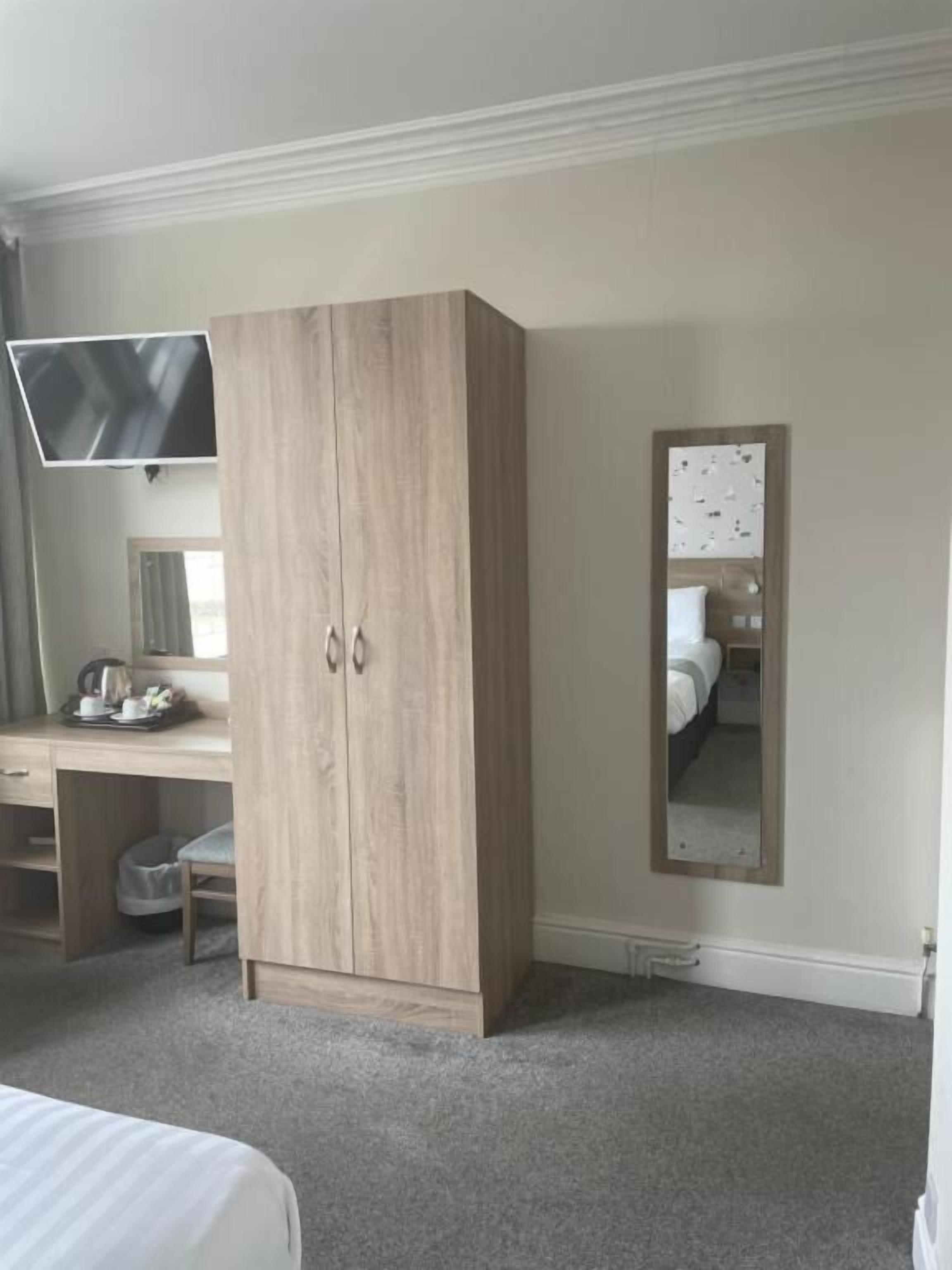 A bedroom with a wooden wardrobe, a desk with a flat-screen TV, a bed, and a mirror on the wall.
