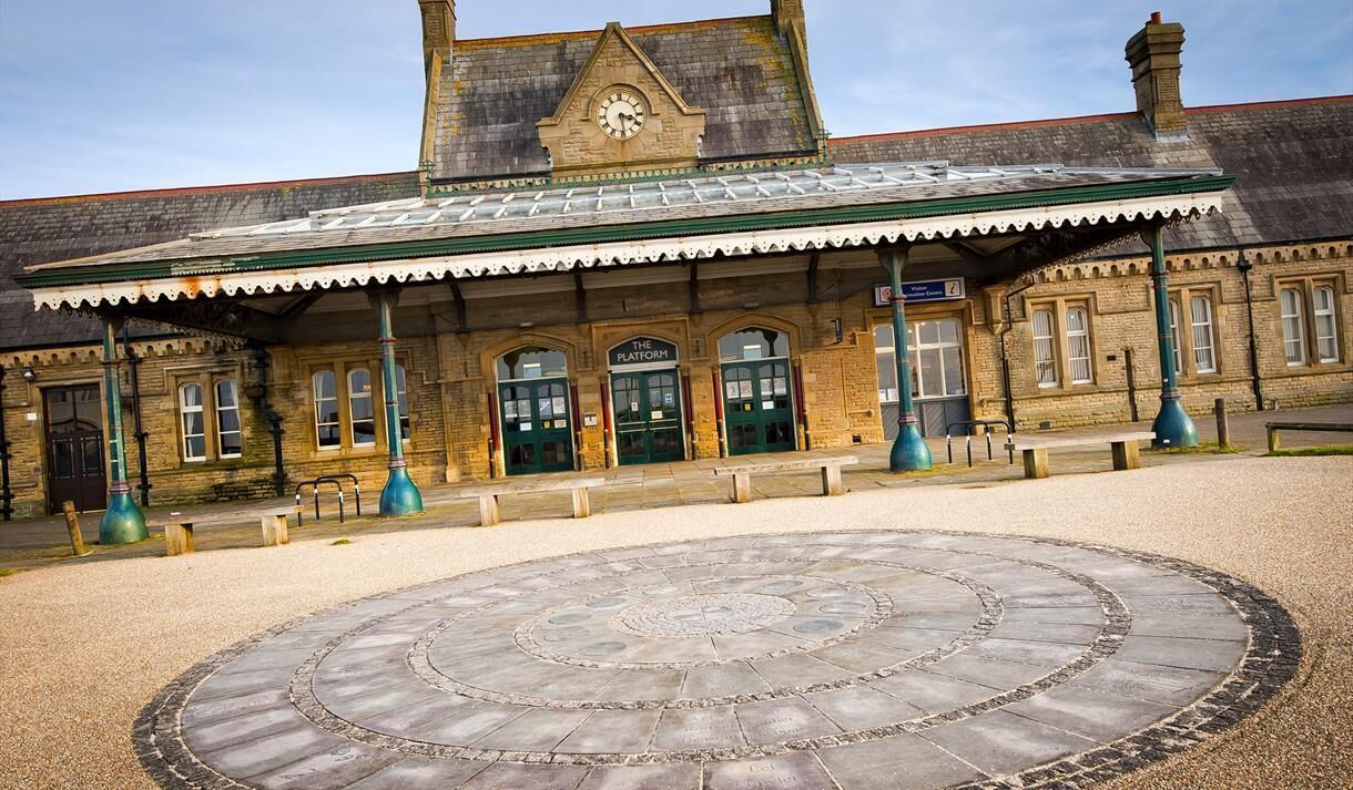 A historic train station with a clock tower, a covered waiting area, and a circular stone pavement in the foreground.